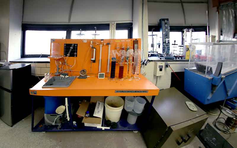 an orange fluid mechanics workbench with many clear cylinders, some with colored liquid, empty containers under the work bench and other lab equipments by the window